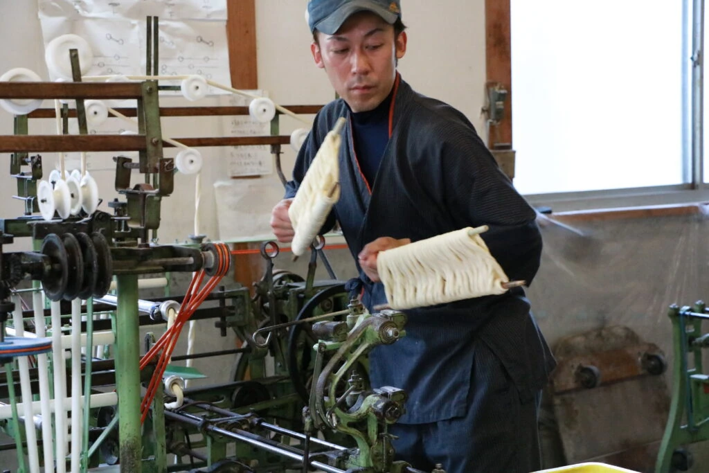 マルカツ製麺所で作務衣姿の職人が製麺機から手延べそうめんを取り上げる様子